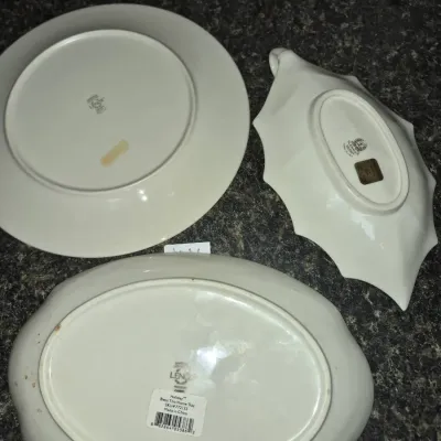 Sale Photo Thumbnail #715: Set of three Lenox fine china serving pieces shown from the underside, including a round plate, a leaf-shaped dish, and an oval 'Bless This Home' tray, all featuring maker's marks.