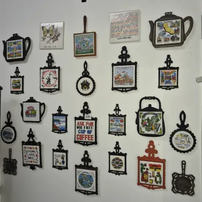 Sale Photo Thumbnail #751: Large collection of vintage decorative trivets and wall plaques featuring travel souvenirs, regional motifs, and ceramic tiles mounted on metal or wooden frames.