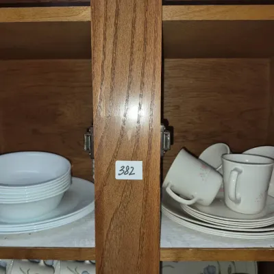 Sale Photo Thumbnail #666: Assorted vintage tableware in a wooden cabinet, including stacked white Corelle-style bowls and plates, alongside floral coffee mugs and saucers.
