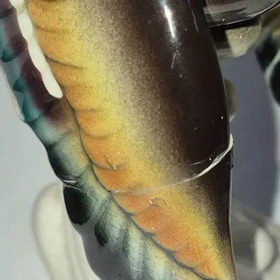 Sale Photo Thumbnail #238: Close-up detail of a glazed ceramic figurine showing a repaired crack along a colorful wing or feather detail.