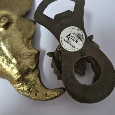 Sale Photo Thumbnail #682: Vintage cast metal or brass bottle opener with an 'Action' manufacturer sticker, showing light surface tarnish.