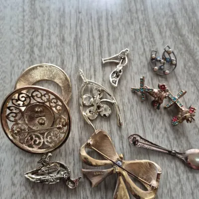 Sale Photo Thumbnail #872: Collection of vintage costume jewelry brooches and pins, including a textured gold-tone bow pin, floral designs, a horseshoe, and a small spoon brooch, shown from the back with visible hardware markings.
