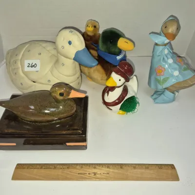 Sale Photo Thumbnail #443: Collection of duck-themed decorative items, including ceramic figurines, plush toys, a covered trinket box, and a wooden goose wearing a raincoat.