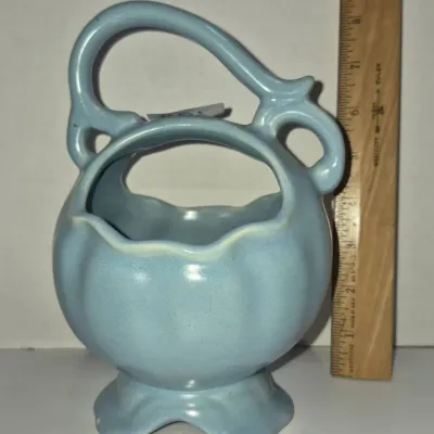 Sale Photo Thumbnail #266: Vintage light blue ceramic pottery basket vase featuring an ornate loop handle and pedestal foot, measuring approximately eight inches tall.