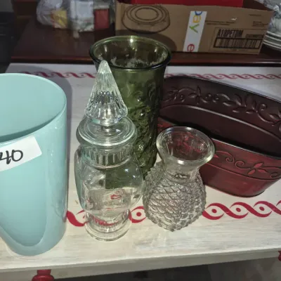Sale Photo Thumbnail #63: Collection of decorative containers, including a light blue ceramic vase, clear glass candy jar with lid, olive green textured glass vase, small cut-glass bud vase, and an embossed reddish-brown metal planter tub.