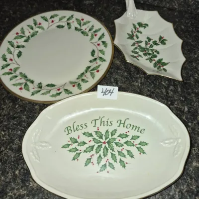 Sale Photo Thumbnail #714: Set of three Lenox-style holiday serveware pieces, including a round dinner plate, a leaf-shaped handle dish, and an oval dish reading "Bless This Home," all decorated with holly foliage and berries.