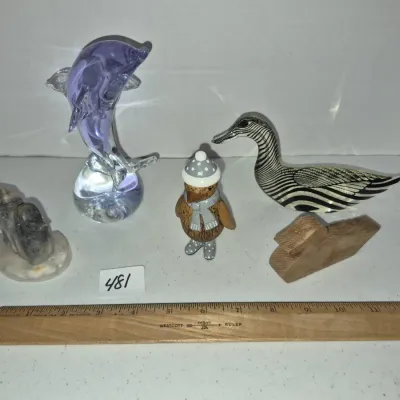 Sale Photo Thumbnail #826: Collection of five decorative animal figurines, including a purple glass dolphin, zebra-patterned art glass duck, carved stone bird, wooden penguin in winter attire, and a small wooden cut-out, shown alongside a ruler for scale.