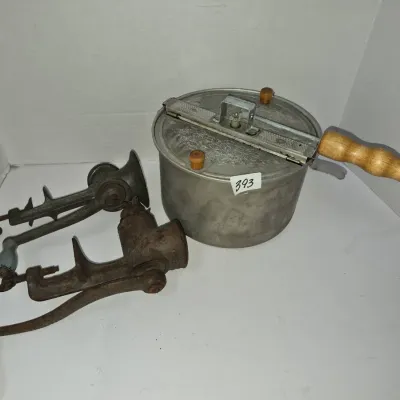 Sale Photo Thumbnail #687: Vintage aluminum stovetop popcorn popper with a wooden hand crank alongside two cast iron hand-crank food grinders.