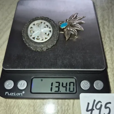 Sale Photo Thumbnail #851: Two vintage sterling silver brooches on a digital scale displaying a weight of 13.40 grams, including one round mother-of-pearl inset piece and one leaf-shaped pin with a turquoise cabochon.
