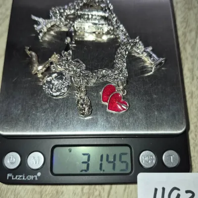 Sale Photo Thumbnail #846: Sterling silver charm bracelet featuring various travel and novelty charms, including red enameled hearts, a pineapple, and an Eiffel Tower, weighing 31.45 grams on a digital scale.