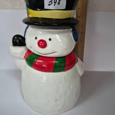 Sale Photo Thumbnail #607: Festive ceramic snowman cookie jar featuring a black top hat, red and green striped scarf, and blue earmuffs, tagged with lot number 348.