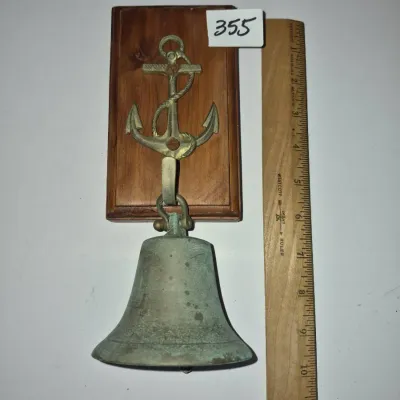 Sale Photo Thumbnail #618: Nautical-themed brass or bronze ship's bell mounted on a wooden backplate, featuring a stylized anchor hook hanger.