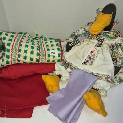 Sale Photo Thumbnail #276: Vintage handmade dressed goose doll with wire-rim glasses alongside a collection of folded linens, including a red-and-green woven throw.
