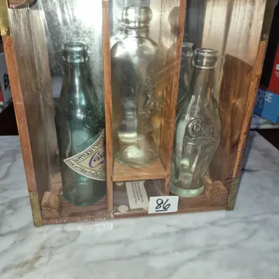 Sale Photo Thumbnail #137: Vintage Coca-Cola commemorative glass bottle collection housed in a wooden display crate with brass accents, sealed in plastic.