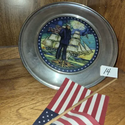Sale Photo Thumbnail #22: Vintage pewter commemorative plate featuring a stained glass-style center illustration of a sailor and tall ships, displayed with a small paper American flag.
