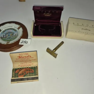 Sale Photo Thumbnail #425: Vintage collectibles including a 1964 New York World's Fair souvenir ashtray with wooden base, a Bulova Excellency watch box set, a safety razor, and a vintage restaurant matchbook.