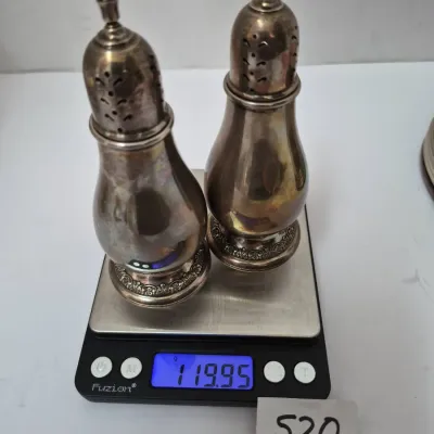 Sale Photo Thumbnail #897: Pair of vintage sterling silver salt and pepper shakers with ornate detailed bases, shown on a digital scale weighing 119.95 grams.