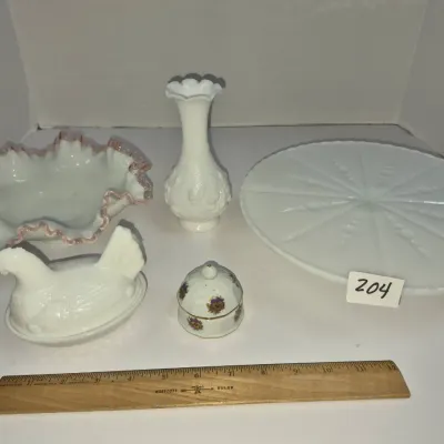 Sale Photo Thumbnail #346: Collection of vintage milk glass tableware including a ruffled bowl with pink trim, a hen on nest covered dish, a bud vase, a cake platter, and a small floral trinket box.
