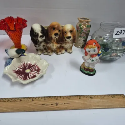 Sale Photo Thumbnail #406: Collection of vintage decorative ceramics and glassware, including a trio of spaniel figurines, a red hobnail ruffle-rim vase, a flower dish with a bird, a girl figurine, and a bowl filled with glass marbles.