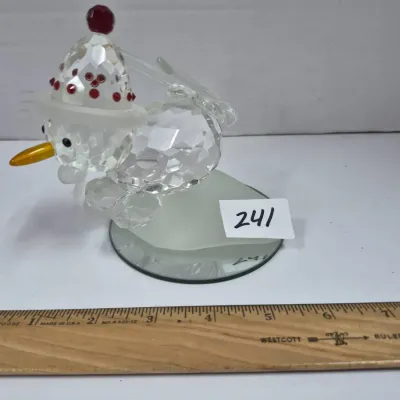 Sale Photo Thumbnail #413: Swarovski crystal snowman figurine with a red-accented winter hat, carrot nose, and mirrored display base, shown alongside a ruler for size reference.