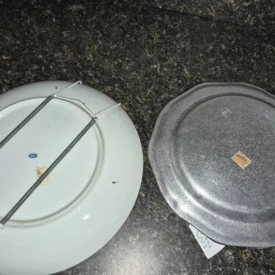 Sale Photo Thumbnail #717: Undersides of two decorative plates, including a white porcelain plate with a wire wall hanger and a stamped pewter-style metal plate.
