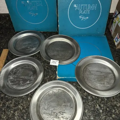 Sale Photo Thumbnail #690: Set of five vintage pewter collector plates featuring etched landscape scenes by Eric Sloane, accompanied by their original blue presentation boxes.