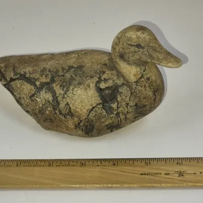 Sale Photo Thumbnail #326: Hand-carved rustic stone or composition duck decoy figurine with a distressed, crackled finish, measuring approximately seven inches long.