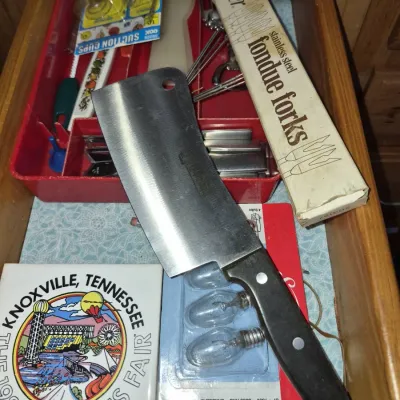 Sale Photo Thumbnail #685: Vintage kitchen drawer containing a meat cleaver, boxed Oster stainless steel fondue forks, a 1982 Knoxville World's Fair souvenir, and various small household items.