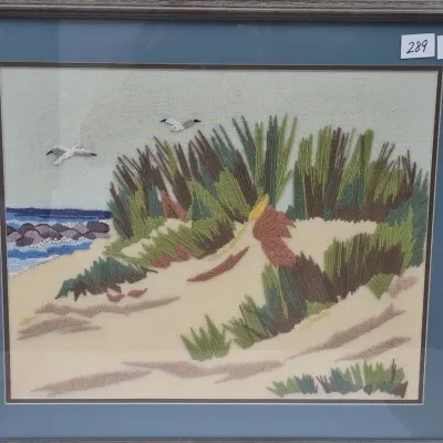 Sale Photo Thumbnail #511: Framed vintage embroidered textile artwork depicting coastal sand dunes with sea grass, flying seagulls, and ocean waves. Presented in a blue mat with a dark wood frame.