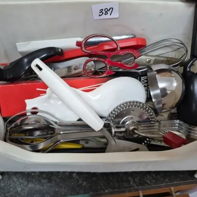 Sale Photo Thumbnail #679: Assorted vintage and modern kitchen utensils stored in a white plastic bin, including a hand rotary egg beater, kitchen shears, and a ceramic spoon rest.