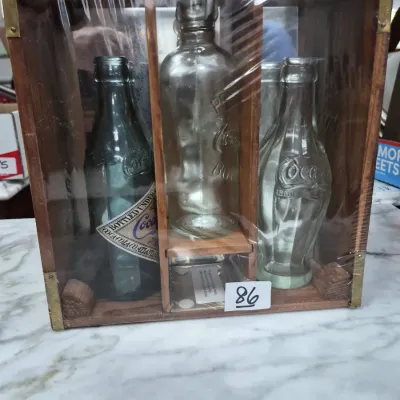Sale Photo Thumbnail #138: Vintage Coca-Cola commemorative glass bottle collection set in a wooden display crate with brass accents, sealed in plastic wrap.