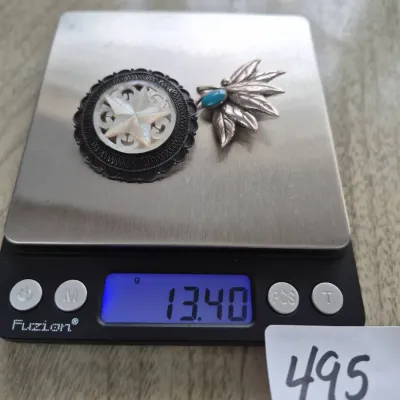Sale Photo Thumbnail #850: Two vintage brooches sitting on a digital scale, including a round mother-of-pearl star motif pin and a leaf-shaped piece with a turquoise accent, weighing a combined 13.40 grams.