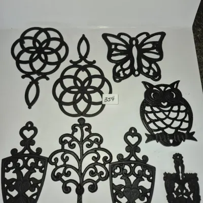 Sale Photo Thumbnail #617: Collection of eight black cast iron trivets featuring butterfly, owl, and traditional openwork scroll designs.