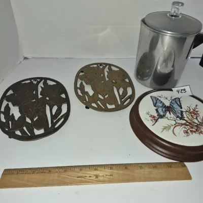 Sale Photo Thumbnail #740: Vintage kitchenware group including two cast brass floral trivets, a wooden trivet with a ceramic butterfly insert, and an aluminum stovetop coffee percolator with a glass knob.