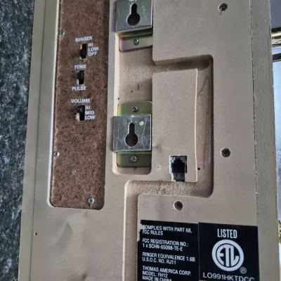 Sale Photo Thumbnail #678: Back panel of a Thomas America Corp vintage-style wall telephone, model FH12, featuring controls for ringer, tone/pulse, and volume alongside mounting hardware.