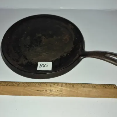 Sale Photo Thumbnail #631: Vintage cast iron round griddle pan with handle, featuring heavy seasoning and patina, displayed next to a wooden ruler for scale.