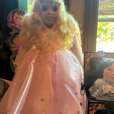 Blonde collector doll wearing a embellished pink ballgown with a jewel brooch on the bodice, displayed alongside a boxed Wizard of Oz doll.
