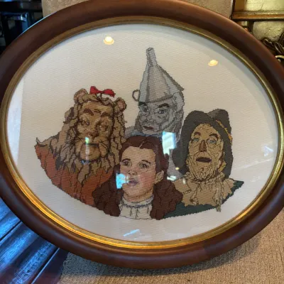 Sale Photo Thumbnail #148: Framed Wizard of Oz cross-stitch artwork, featuring Dorothy, the Cowardly Lion, the Tin Man, and the Scarecrow in a dark wood oval frame with gold inner trim.