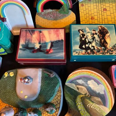 Sale Photo Thumbnail #124: Collection of Wizard of Oz collectibles, including printed trinket boxes, a hand-painted wooden paddle, rainbow figurines, and a decorative display plate.