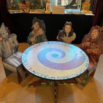 Sale Photo Thumbnail #152: The Wizard of Oz themed table set with cut-out character chairs depicting the Tin Man, Scarecrow, Dorothy, and Cowardly Lion, accompanied by a collection of vintage Oz memorabilia in the background.