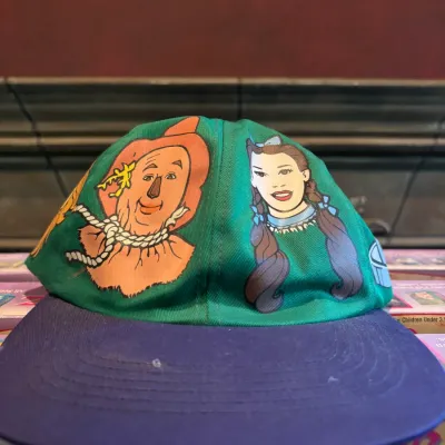Sale Photo Thumbnail #115: Vintage Wizard of Oz baseball cap featuring printed illustrations of the Scarecrow and Dorothy on a green crown with a purple brim.