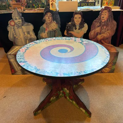 Custom-painted Wizard of Oz themed table featuring a spiral yellow brick road top and floral wooden pedestal base, displayed alongside...
