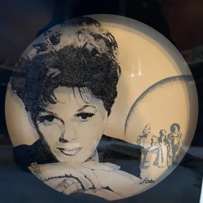 Framed circular Judy Garland artwork print featuring a stippled portrait with a tiny Wizard of Oz silhouette scene in the background,...