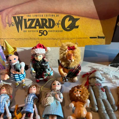 Sale Photo Thumbnail #132: Collection of vintage Wizard of Oz Christmas ornaments featuring characters like Dorothy, the Cowardly Lion, and the Tin Man, displayed in front of a 50th Anniversary poster.
