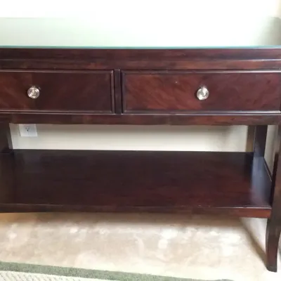 Dark mahogany-finished wooden console table featuring two drawers with round silver-tone knobs, a lower open shelf, and a glass tabletop...