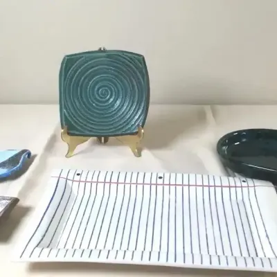 Collection of artisan studio pottery items, including a rectangular platter painted to resemble notebook paper, a teal swirled square...