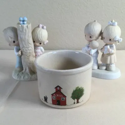 Small ceramic schoolhouse motif crock alongside two Precious Moments porcelain figurines.