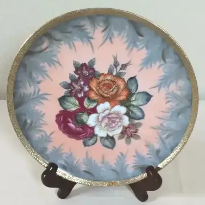 Decorative porcelain display plate featuring a hand-painted floral bouquet on a pink and blue background with a gold-toned textured rim.