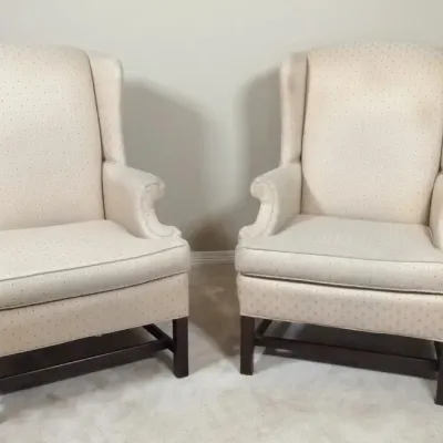 Pair of traditional wingback armchairs upholstered in cream fabric with subtle dot patterning and dark wood stretchered legs.