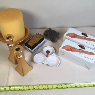 Collection of home decor items including a pair of brass candlesticks, a large pillar candle on a dish, boxed napkin ring sets, and a...